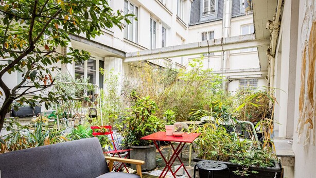 Ein Innenhof mit einer Holzterrasse, einem grauen Sofa, einem roten Tisch und verschiedenen Blumentöpfen.