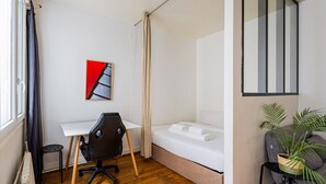 Ein kleines Zimmer mit Bett, Schreibtisch, Stuhl, Heizkörper und einem Topfpflanzen.