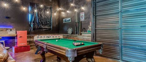 Een pooltafel in een ruimte met een dartbord, een bord waarop 'Farnsworth' staat en een gesloten garage deur.