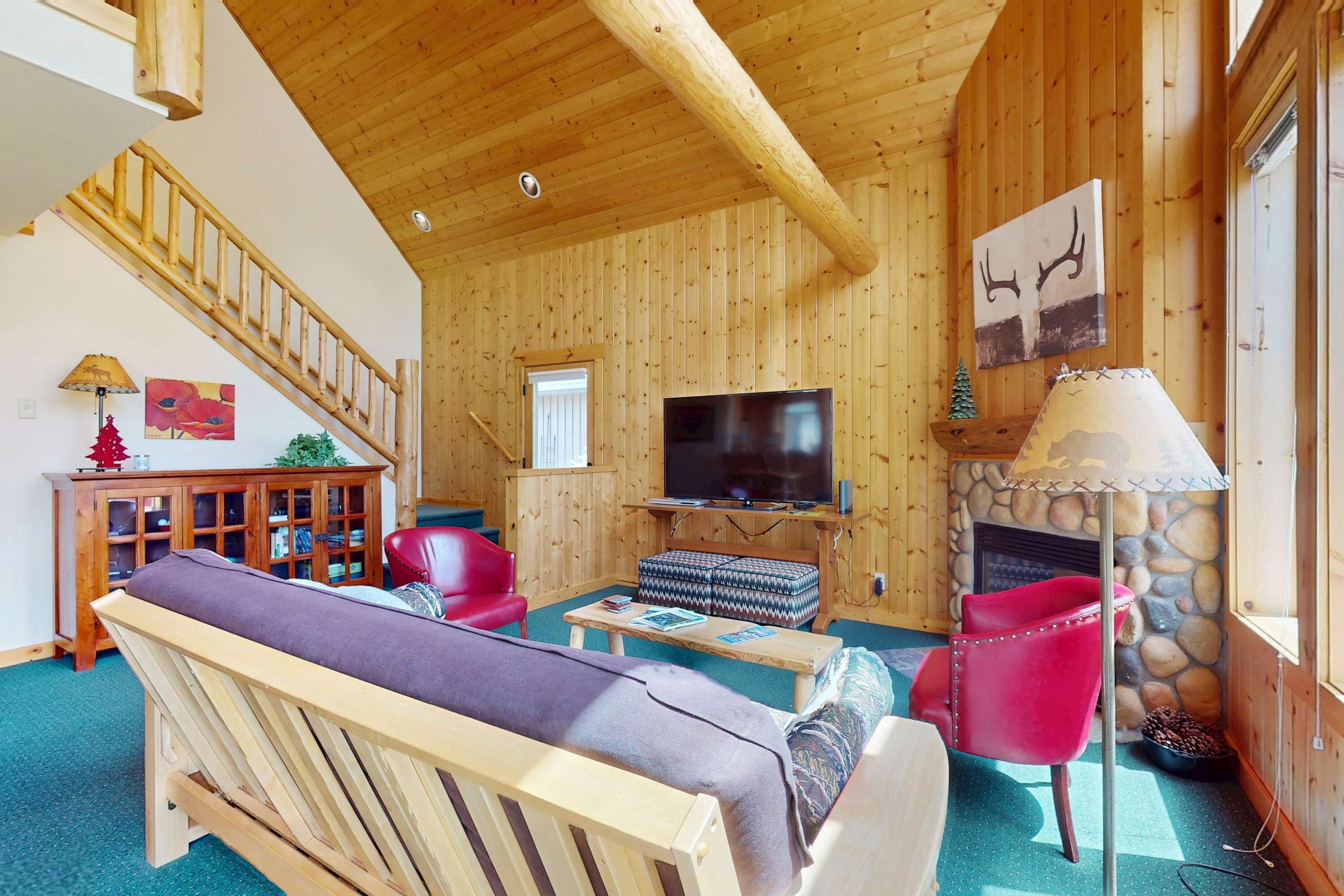 A wooden interior with a TV, red chairs, a sofa, a fireplace, and a staircase.