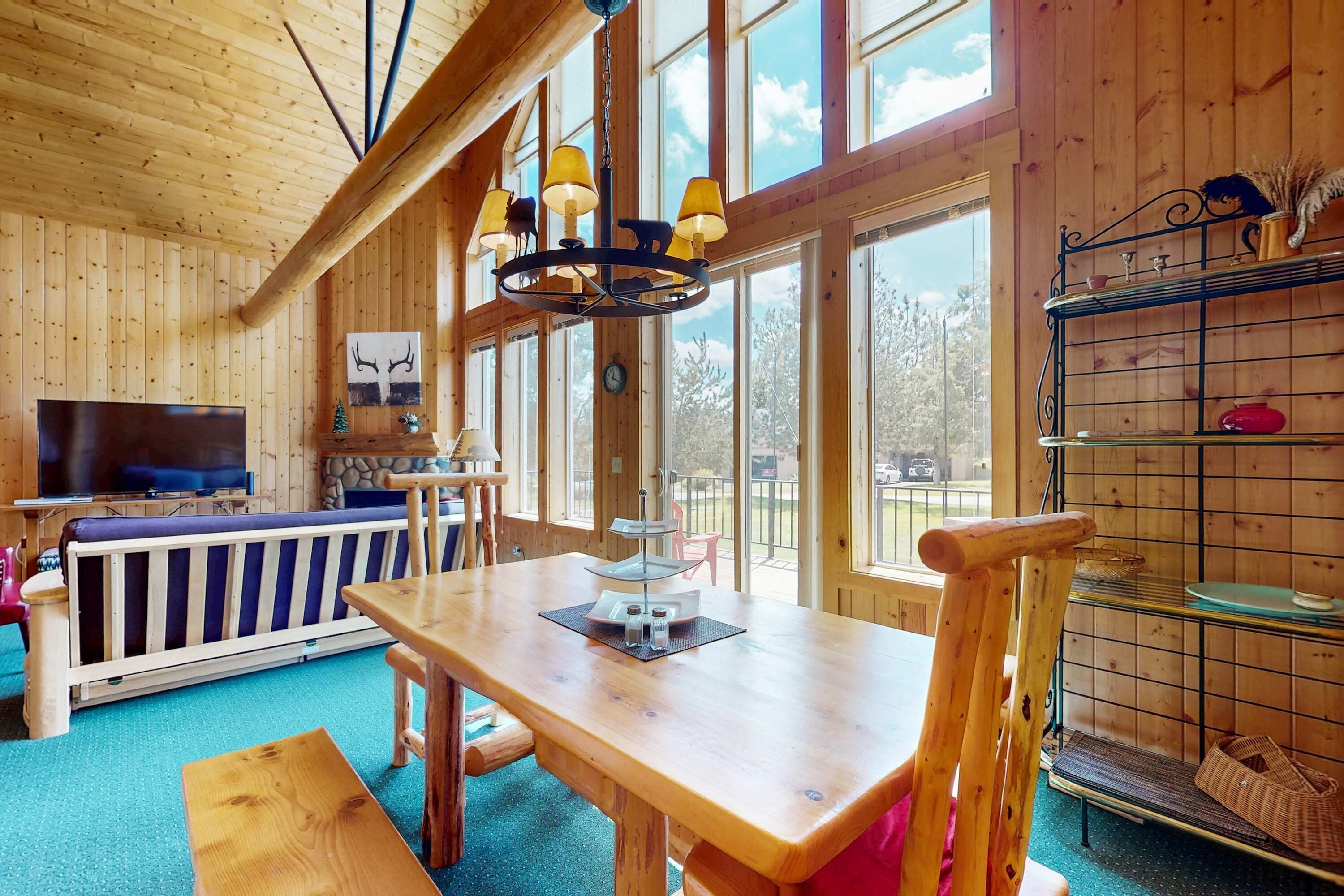 A wooden interior with a dining table, chairs, a TV, and large windows.