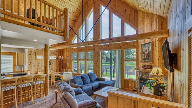 A wooden cabin interior with a dining area, seating, a fireplace, and large windows.