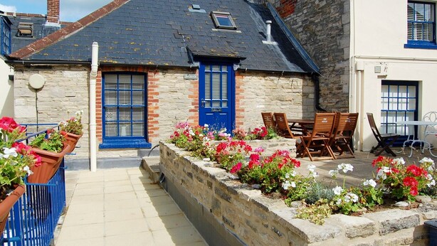 Un bâtiment en pierre avec une porte bleue, des places assises à l’extérieur et des parterres de fleurs.
