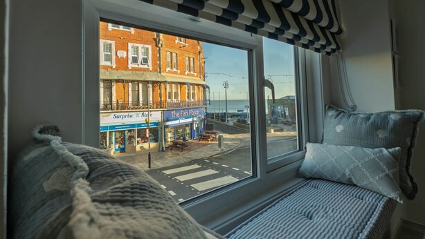 Une chambre dotée d’une grande fenêtre donnant sur la rue, d’un lit recouvert d’une couverture à rayures et d’un oreiller orné d’un motif en forme de coquillage.