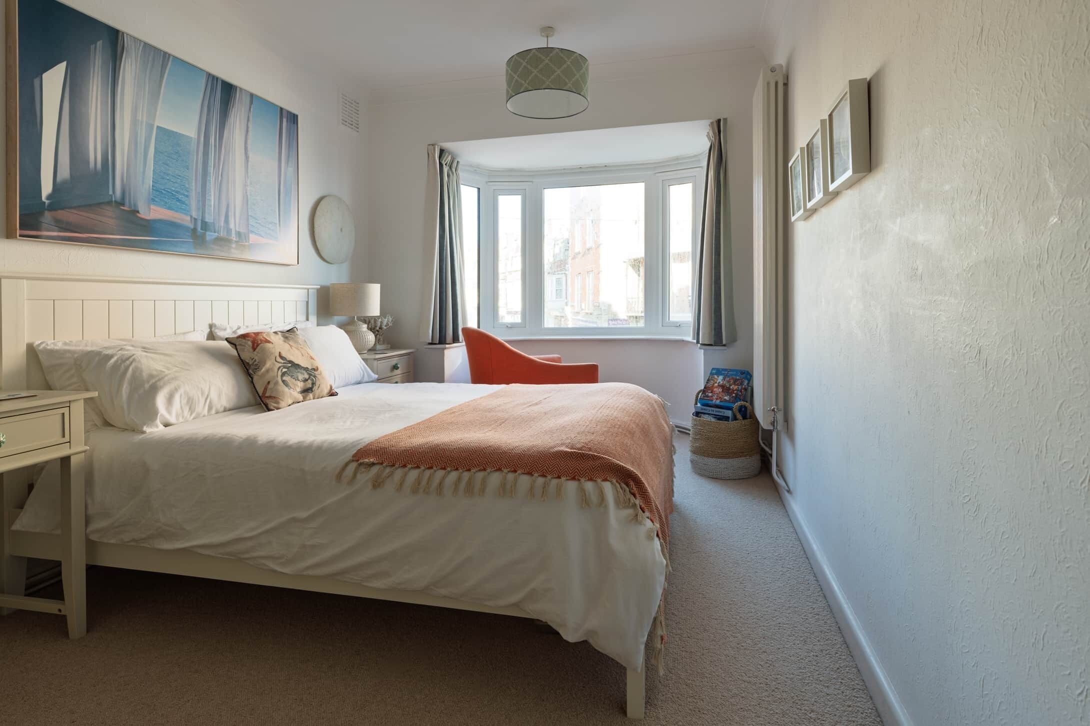 A bedroom with a bed, a nightstand, a lamp, a window with curtains, a painting on the wall, and a view of the street outside.