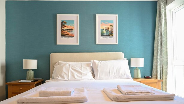 A hotel room with a bed, two framed pictures on the wall, two bedside lamps, and a window with curtains. - Perfect Family Base: Spacious Seaside Apartment with Parking (Swanage)