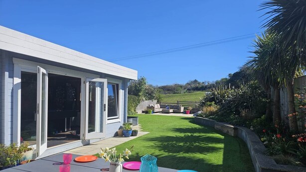 A patio with a table set for dining, a house with a sliding glass door, and a well-maintained lawn. - Spacious Countryside Home with Games Room & Large Garden Near Swanage (Langton Matravers)