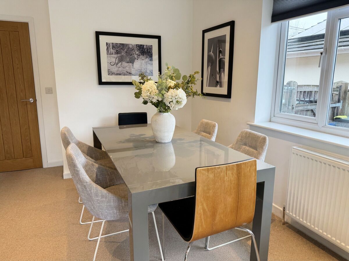 A dining area with a glass table, grey and wooden chairs, a white vase with flowers, and framed pictures on the wall.