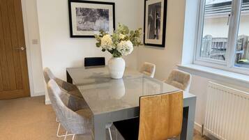 A dining area with a glass table, grey and wooden chairs, a white vase with flowers, and framed pictures on the wall.