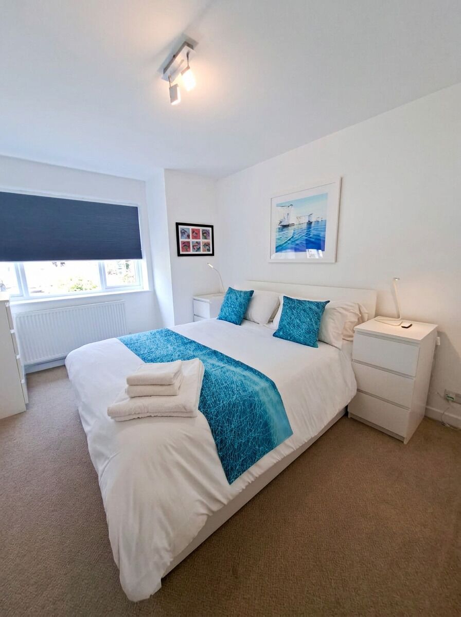A bedroom with a large bed, a bedside table, a nightstand, a window with blinds, and a wall-mounted picture.