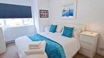 A bedroom with a large bed, a bedside table, a nightstand, a window with blinds, and a wall-mounted picture.