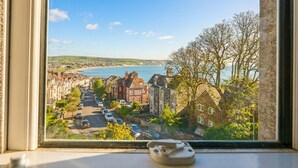 A coastal town view from a window, including a street with parked cars, residential buildings, and a body of water.
