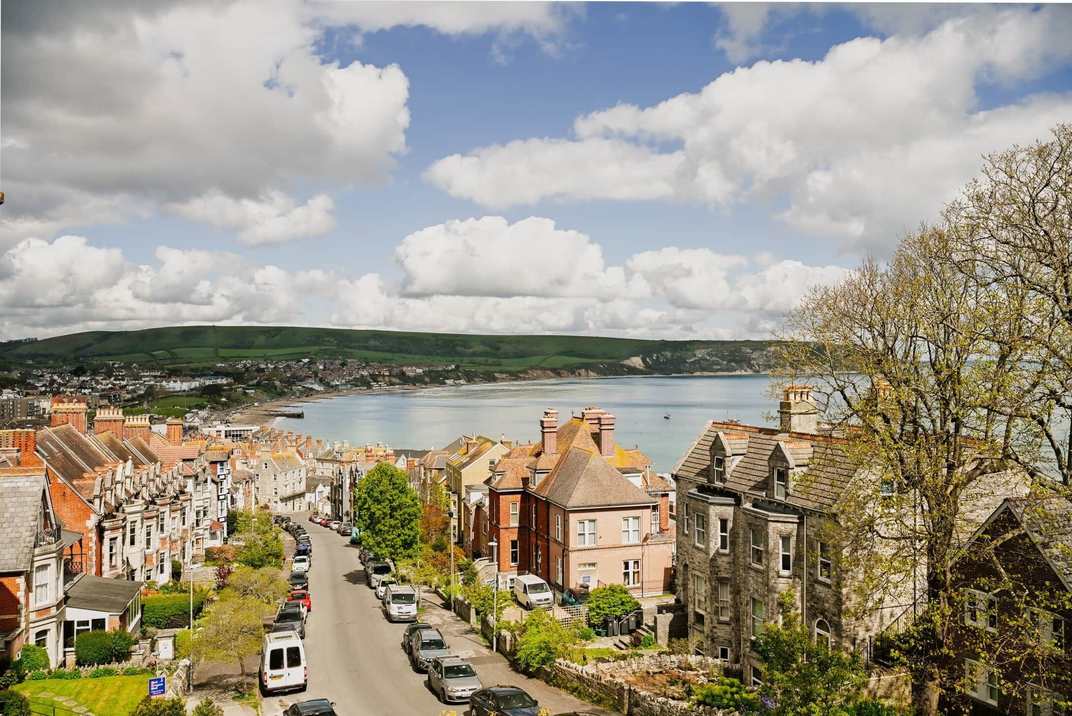A coastal town with terraced houses, a road lined with parked cars, and a body of water in the background.