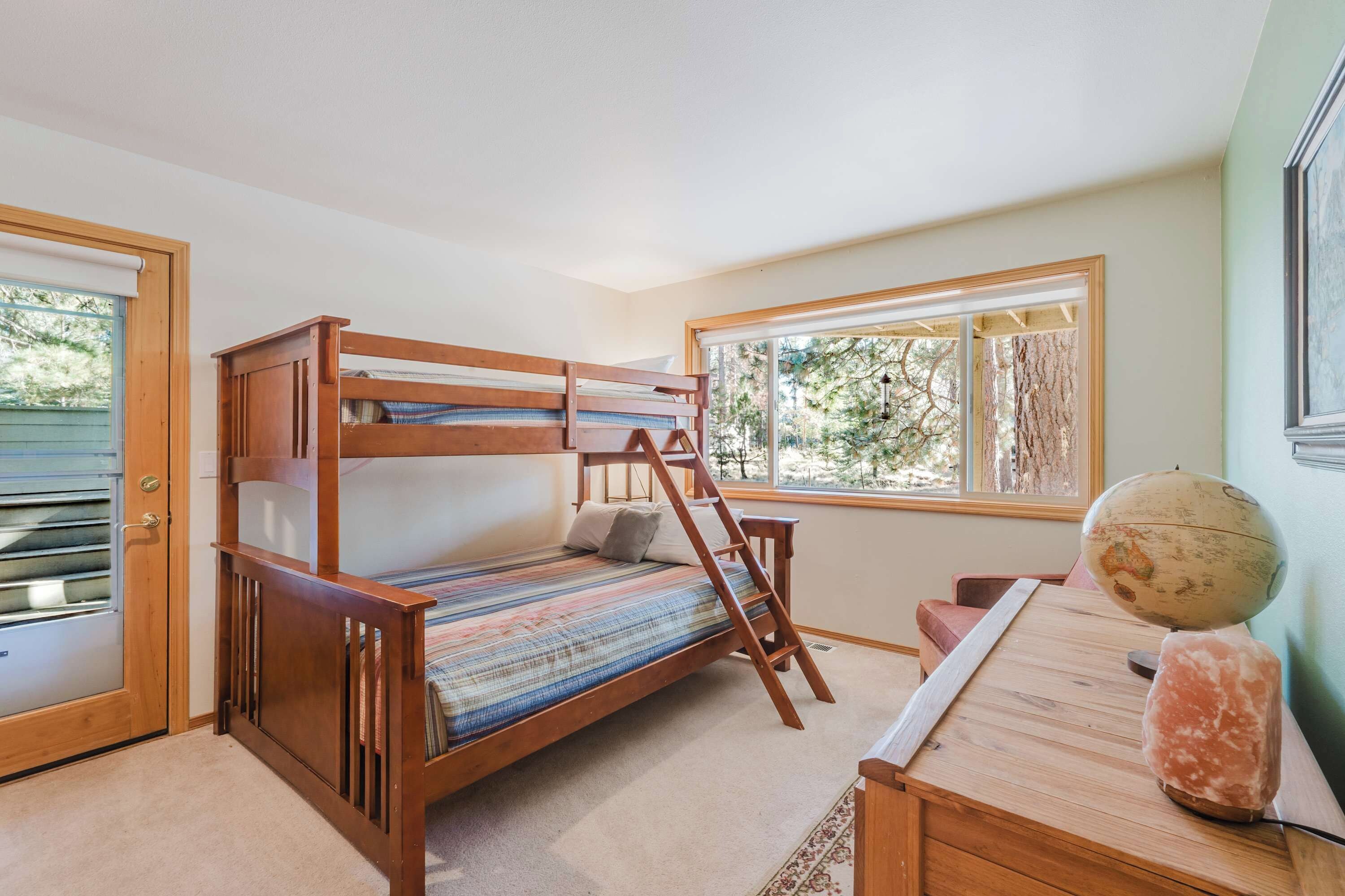 A bunk bed with a ladder, a desk with a globe and a salt lamp, and a window with a view of trees.
