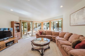 A living room with a television, a bookshelf, a brown sofa, a glass coffee table, and large windows. - Elegant home with pool, private hot tub, game room, deck & SHARC passes (Sunriver)