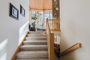 A carpeted staircase with wooden handrails leading to a living area with a view of trees through large windows. - Elegant home with pool, private hot tub, game room, deck & SHARC passes (Sunriver)