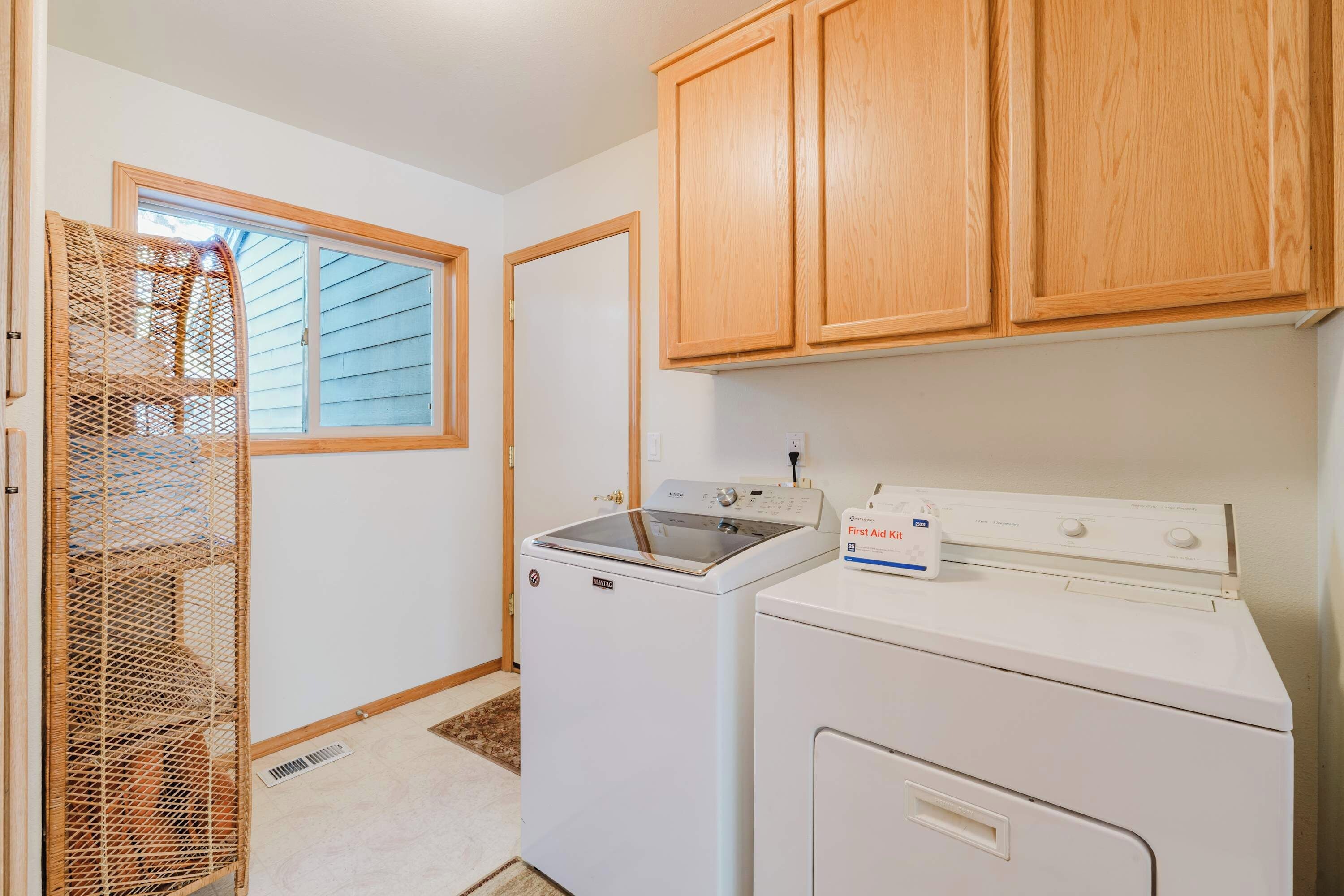 A laundry room with a washing machine, dryer, wooden cabinets, a window, and a wicker basket.
