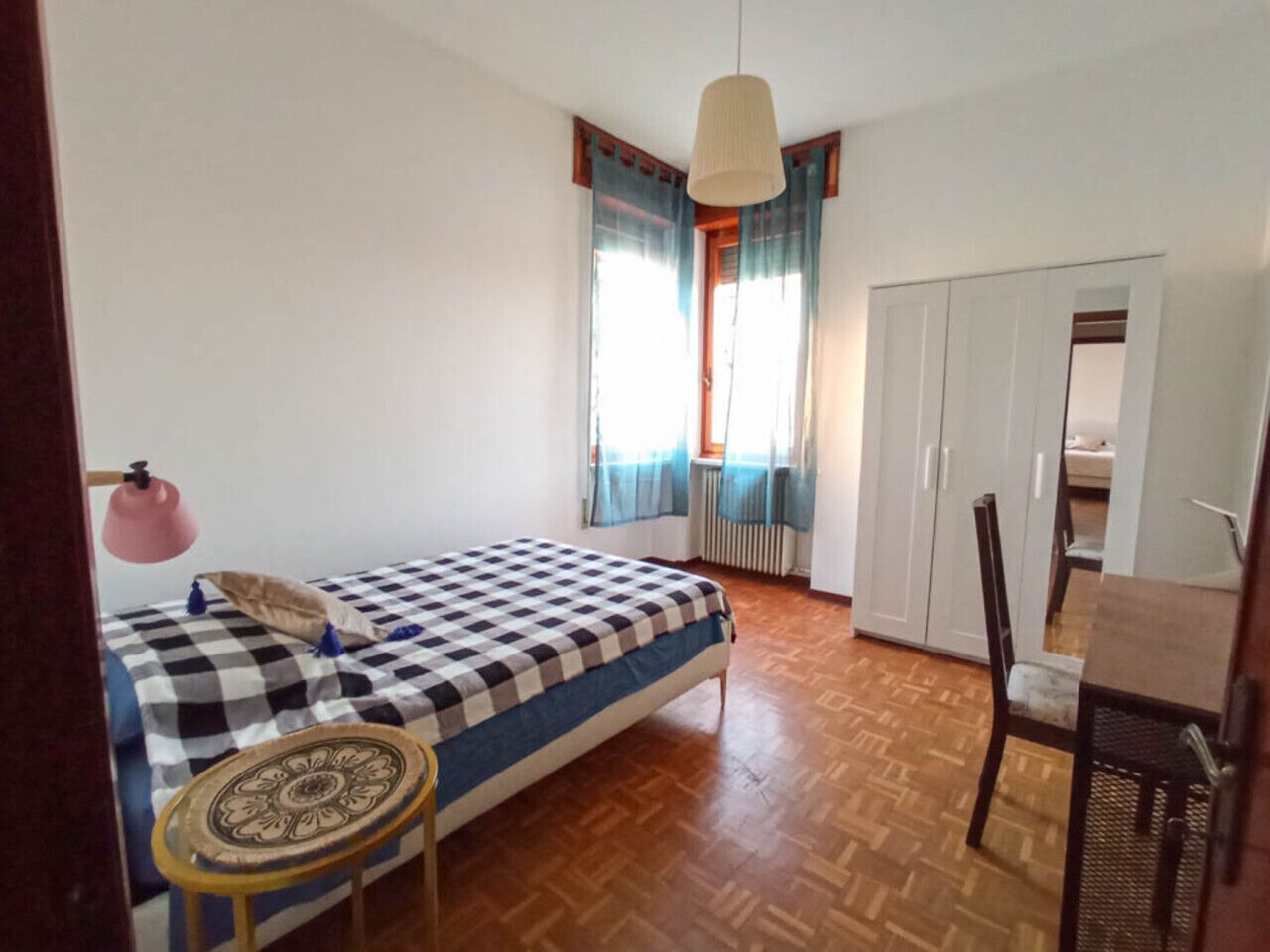 Ein Schlafzimmer mit einem Bett, einem kleinen Tisch, einem Stuhl, einem Schrank und einem Fenster mit Vorhängen.