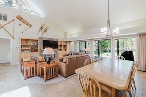 A spacious living room with a large wooden dining table, a sofa, and a TV. - Beautiful Home withPrivate Hot Tub, Deck, Gas Fireplace, and SHARC Passes (Sunriver)