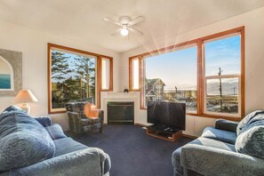 A living room with a fireplace, large windows, a ceiling fan, and a television. - Ocean view home with WiFi, gas fireplaces, private washer&dryer, and foosball (Seaside)