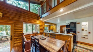 A log cabin interior with a dining area, kitchen, and a balcony. - Spacious riverfront lodge with beach access, private hot tub & forest views (Washougal)
