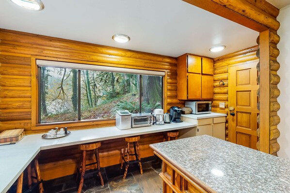 A log cabin kitchen with a large window, wooden cabinets, a granite countertop, and a bar stool.
