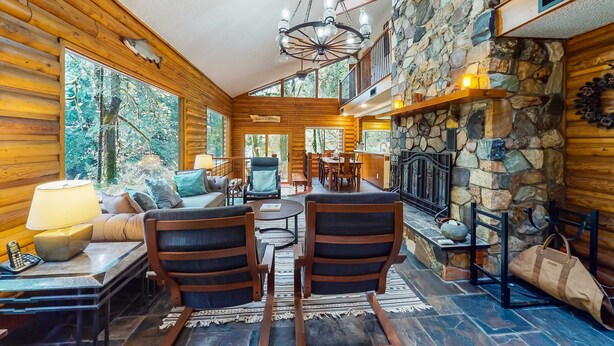 A log cabin interior with a stone fireplace, wooden walls, and a dining area. - Spacious riverfront lodge with beach access, private hot tub & forest views (Washougal)