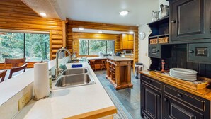 A modern kitchen with wooden cabinetry, a central island, stainless steel sink, and a view of the forest. - Spacious riverfront lodge with beach access, private hot tub & forest views (Washougal)