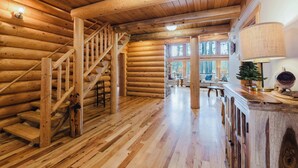 Interior de una cabaña de troncos con una escalera de madera, una mesa de comedor de madera y un amplio ventanal.