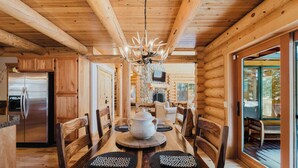 Interior de una cabaña de madera con una mesa de comedor, sillas, una araña, un refrigerador y una chimenea de piedra.