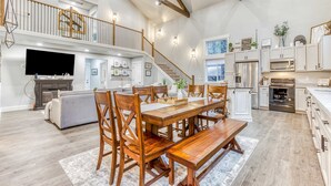 A spacious living area with a dining table, wooden chairs, a sofa, and a staircase leading to an upper level. - Inviting home with fireplace, sauna, full kitchen, & W&D (Welches)