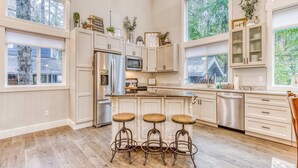A kitchen with wooden flooring, white cabinetry, a central island with stools, and large windows. - Inviting home with fireplace, sauna, full kitchen, & W&D (Welches)