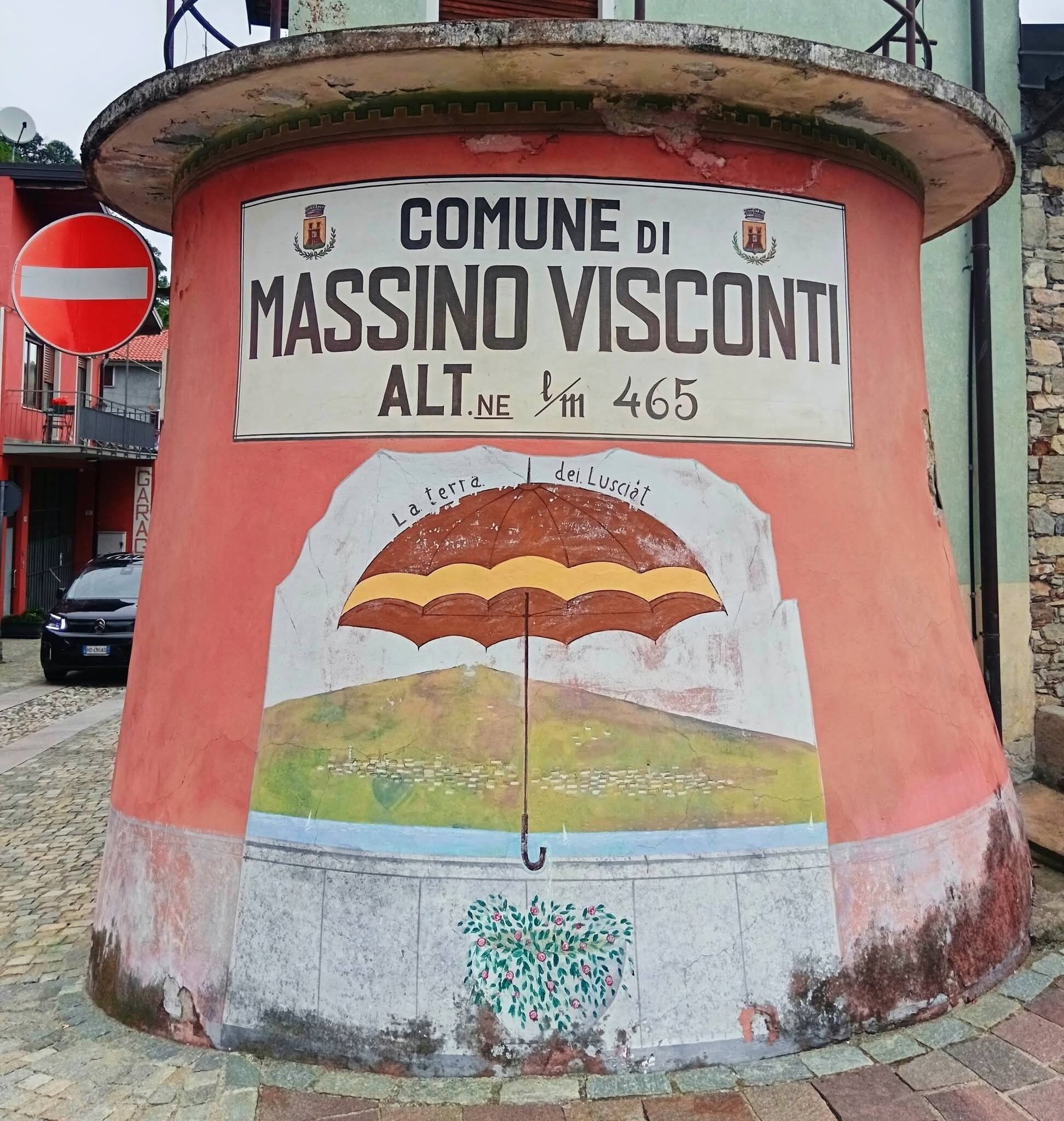 Pyöreä, sään kuluttama kyltti, jossa lukee ”COMUNE DI MASSINO VISCONTI” ja puhelinnumero.
