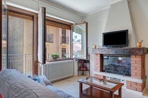 Una sala de estar con un sofá, una mesa de centro de vidrio, una chimenea, un televisor y un balcón con vistas a edificios.