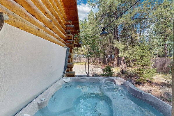 A hot tub with a wooden deck and a view of a forest. - Cozy log cabin with wrap-around porch, Hot tub Loft space, Washer & Dryer (Bend)