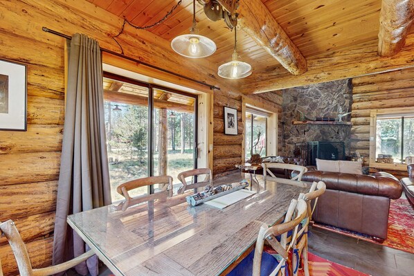 A log cabin interior with a dining table, chairs, a leather sofa, a fireplace, and large windows. - Cozy log cabin with wrap-around porch, Hot tub Loft space, Washer & Dryer (Bend)