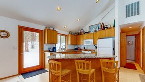 A kitchen with wooden cabinets, a microwave, a refrigerator, and a bar area with three chairs. - Family-Friendly Sunriver Home with Private Hot Tub, Shared Pool, WiFi, SHARC passes (Sunriver)