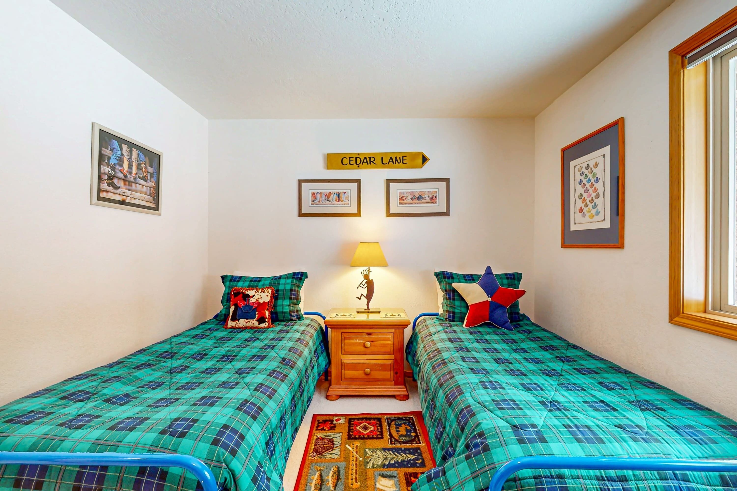 A room with two beds, a nightstand with a lamp, a sign reading 'CEDAR LANE,' and framed pictures on the walls.