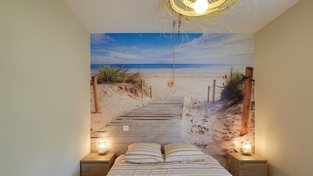 A bedroom with a beach mural, a wooden bed, two bedside tables with lamps, and a hanging light fixture.