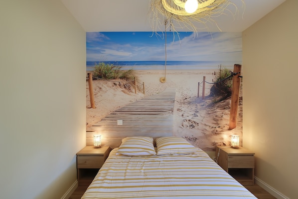 Ein Schlafzimmer mit Wandgemälde vom Strand, einem hölzernen Bett, zwei Nachttischen mit Lampen und einer Pendelleuchte.