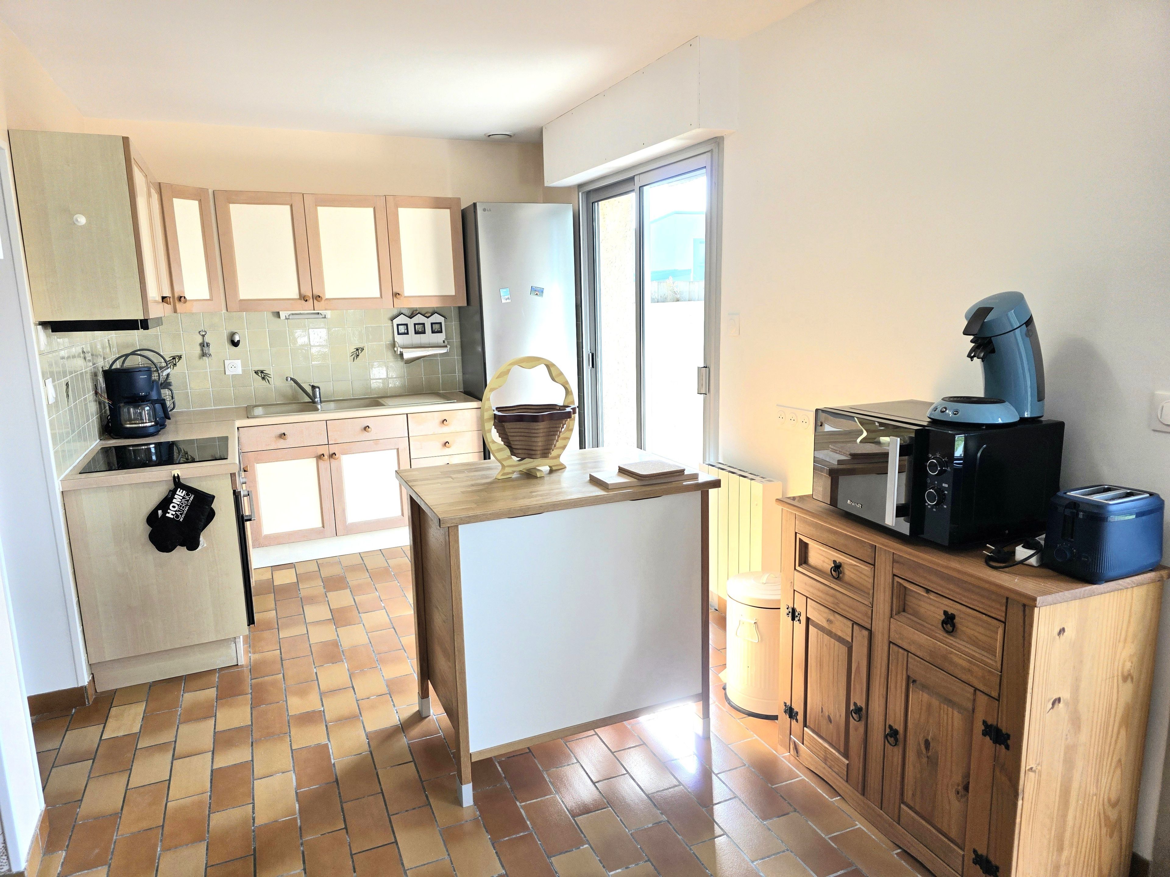 Cocina compacta con cafetera, microondas y tostadora sobre la encimera, armarios de madera y salpicadero de azulejos.