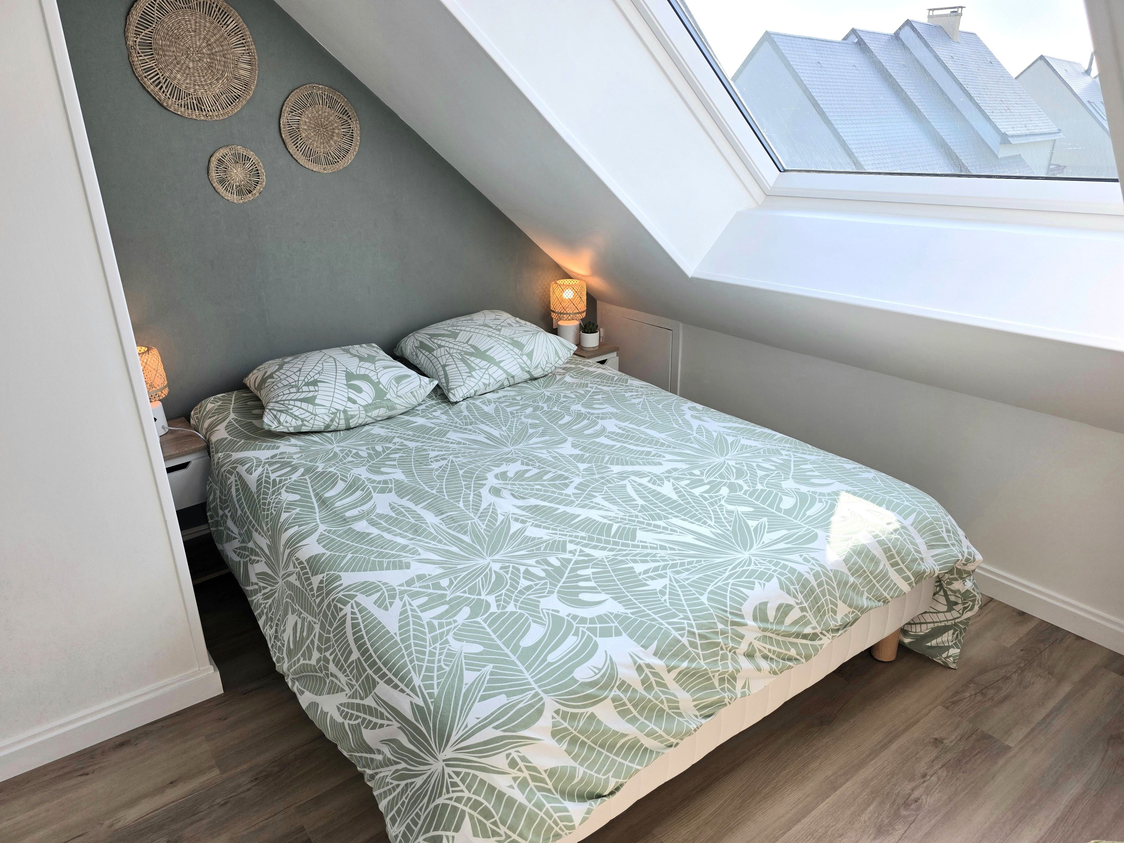 A bedroom with a sloped ceiling, a bed with leaf-patterned bedding, a bedside lamp, and a view of buildings through a skylight.