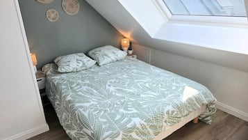 A bedroom with a sloped ceiling, a bed with leaf-patterned bedding, a bedside lamp, and a view of buildings through a skylight.