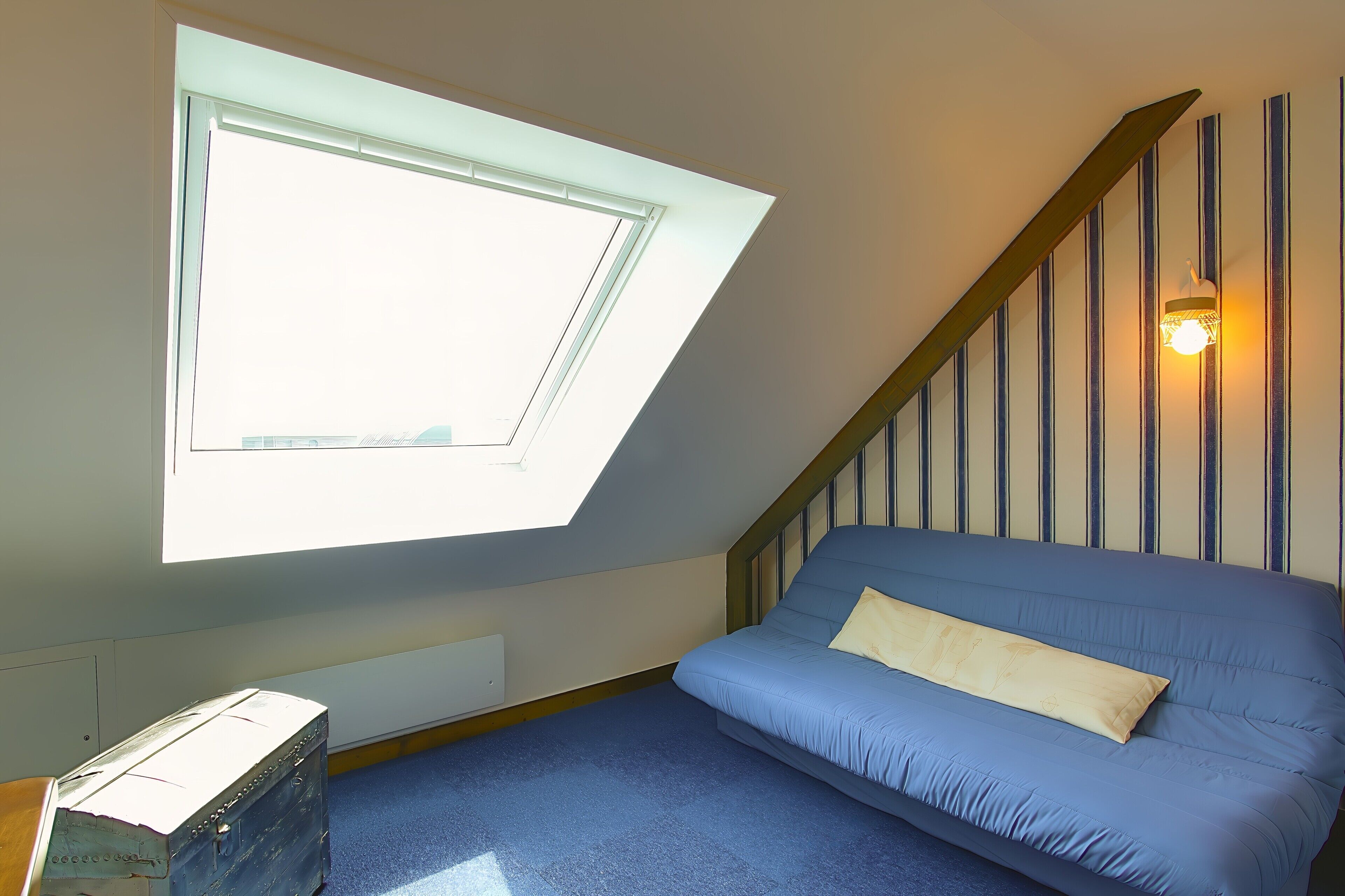 Een kamer met een schuine kap, een dakraam, een blauwe bank, een houten kist en een wandlamp.