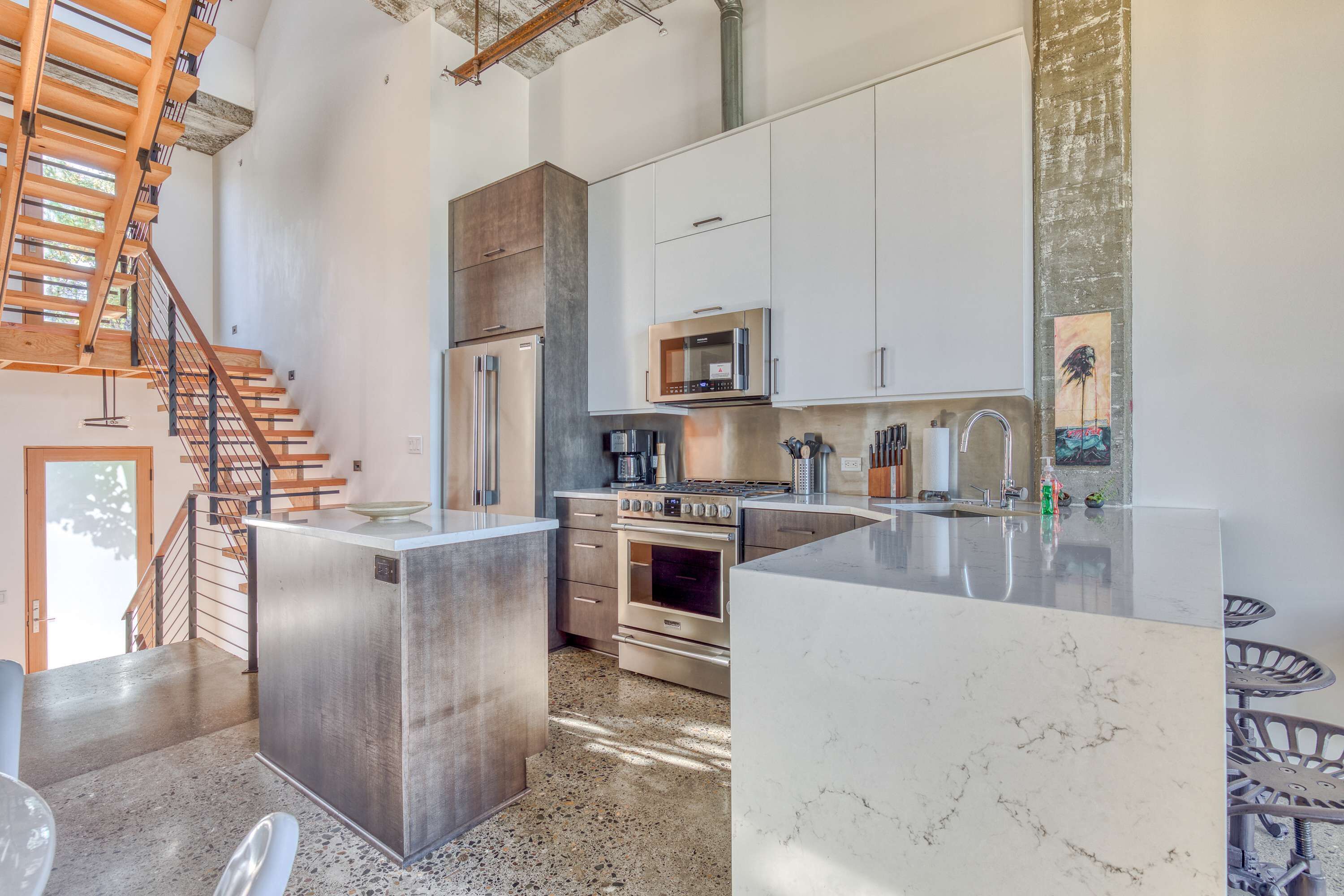 A modern kitchen with stainless steel appliances, a marble island, and a staircase.