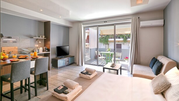 Quarto de hotel moderno com cama, escrivaninha com televisão, varanda com mesa e cadeiras, e vista para o exterior.