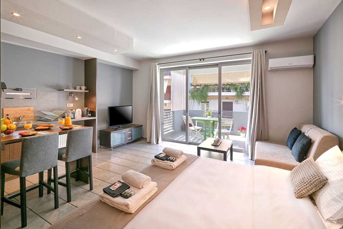 A modern hotel room with a bed, a desk with a TV, a balcony with a table and chairs, and a view of the outside.