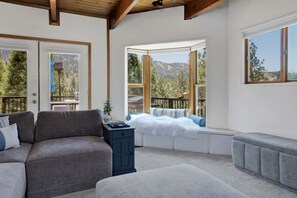 A cozy room with a sofa, ottoman, and a small table. Large windows offer a view of the outdoors. - Big Bear Cabin w/ Hot Tub, Views, Deck & Firepit (Big Bear Lake)