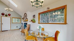 Un coin repas avec une table ronde en verre, des chaises jaunes, un lustre, une grande fenêtre donnant sur les arbres et une cheminée avec une manteau.