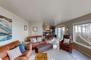 A spacious living room with a large window, a brown leather sofa, a brown leather armchair, and a wooden coffee table. - Spacious home with stunning ocean views, hot tub, shuffleboard, air hockey, WiFi (Waldport)